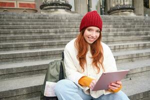 lindo jovem moderno menina com vermelho cabelo, detém digital tábua, senta em escadas perto museu e conecta público Internet, envia mensagem em gadget aplicativo foto