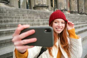 jovem ruiva turista leva selfie dentro frente do museu em escadaria, detém Smartphone e parece às Móvel Câmera, faz foto do ela mesma com telefone