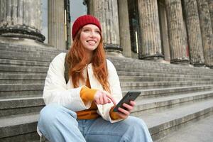 retrato do jovem urbano menina dentro vermelho chapéu, senta em escadas perto museu, detém Móvel telefone, conecta para público Wi-fi e surfa líquido, usa Smartphone apps foto