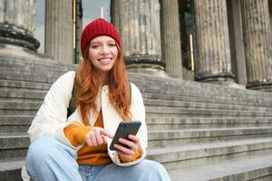 retrato do jovem urbano menina dentro vermelho chapéu, senta em escadas perto museu, detém Móvel telefone, conecta para público Wi-fi e surfa líquido, usa Smartphone apps foto