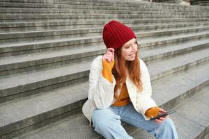 retrato do jovem urbano menina dentro vermelho chapéu, senta em escadas perto museu, detém Móvel telefone, conecta para público Wi-fi e surfa líquido, usa Smartphone apps foto