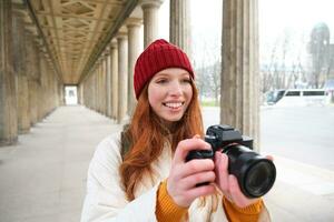 jovem ruiva fêmea fotógrafo, faz estilo de vida tiroteio dentro cidade Centro, leva fotos e sorrisos, parece para perfeito tomada, faz cenário