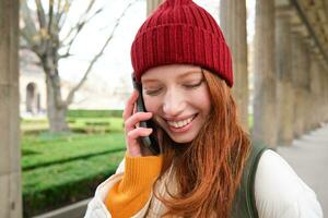 retrato do ruiva europeu menina dentro vermelho chapéu, faz uma telefone chamar, anda em dentro cidade e fala para amigo em Smartphone foto