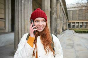 sorridente ruiva fêmea turista fala em Móvel telefone e anda em por aí cidade. feliz aluna dentro vermelho chapéu chamadas amigo, carrinhos em rua e usa Smartphone foto