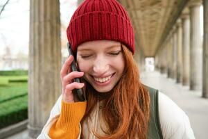 retrato do ruiva europeu menina dentro vermelho chapéu, faz uma telefone chamar, anda em dentro cidade e fala para amigo em Smartphone foto