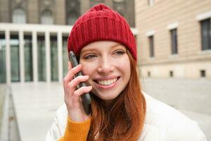 Móvel conexão e pessoas conceito. feliz ruiva mulher dentro chapéu, fala em Móvel telefone, fazer Telefone chamar, usando aplicativo para ligar no exterior foto