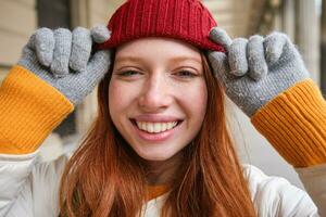 Tiros na Cabeça do feliz ruiva menina com sardas, desgasta vermelho chapéu e luvas dentro inverno, anda em por aí cidade em frio clima e sorrisos foto