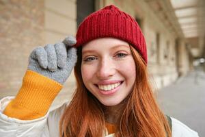 retrato do jovem ruiva mulher dentro tricotado chapéu e luvas, sorrisos e parece aparte, anda em por aí cidade dentro inverno foto