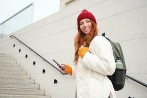 ruiva garota, jovem mulher turista com mochila, detém Smartphone, parece para rota em Móvel aplicativo, pesquisas para hotel em telefone mapa, sorrisos alegremente foto