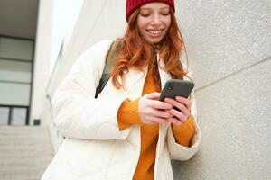 ruiva garota, jovem mulher turista com mochila, detém Smartphone, parece para rota em Móvel aplicativo, pesquisas para hotel em telefone mapa, sorrisos alegremente foto