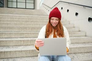 sorridente ruiva garota, aluna senta em escadas ao ar livre e usa computador portátil, conecta para público Wi-fi dentro cidade e trabalho em projeto, usa Internet em computador foto