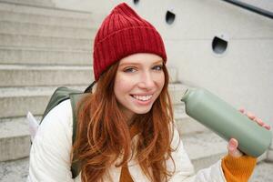 feliz ruiva mulher, turista com garrafa térmica, bebidas dela quente chá, café a partir de viagem frasco, testes de descanso durante dela viajando dentro cidade e passeios turísticos foto