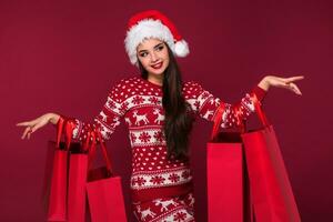 jovem bonita mulher ou menina com grandes lindo cabelo dentro santa chapéu e Novo anos vestir segurando vermelho pacote em vermelho estúdio fundo foto