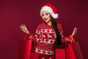 jovem bonita mulher ou menina com grandes lindo cabelo dentro santa chapéu e Novo anos vestir segurando vermelho pacote em vermelho estúdio fundo foto