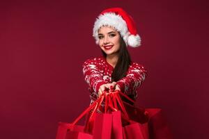 jovem bonita mulher ou menina com grandes lindo cabelo dentro santa chapéu e Novo anos vestir segurando vermelho pacote em vermelho estúdio fundo foto