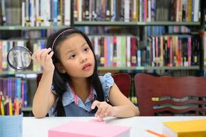 menina asiática segurando a lupa e pensando na biblioteca da escola. conceito de educação e aprendizagem foto