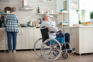 Desativado homem sentado dentro cadeira de rodas dentro cozinha olhando através janela enquanto esposa é preparando café da manhã. inválido, pensionista, deficiente, paralisia. foto