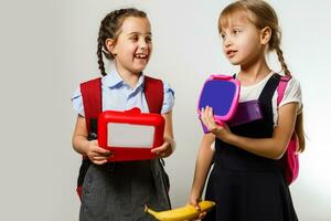 alunos do primário escola com lancheiras dentro mãos. meninas com mochilas estão comendo fruta. começando do aulas. primeiro dia do cair. foto