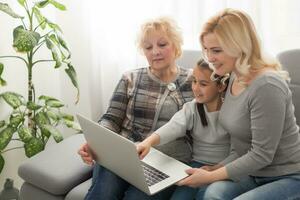 feliz três gerações do mulheres sentar relaxar em sofá rir assistindo engraçado vídeo em computador portátil, sorridente positivo fêmeas avó, mãe e filha ter Diversão descansar em sofá apreciar filme em computador foto