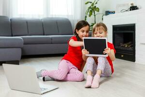 pequeno meninas jogando em uma tábua Informática dispositivo sentado em a chão foto
