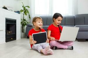 pequeno meninas jogando em uma tábua Informática dispositivo sentado em a chão foto