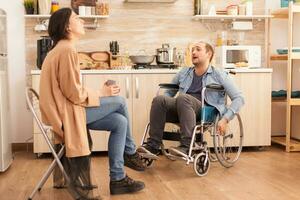 Bravo homem dentro cadeira de rodas Porque do emocional dificuldades com esposa dentro cozinha. Desativado marido discutindo com cônjuge. foto