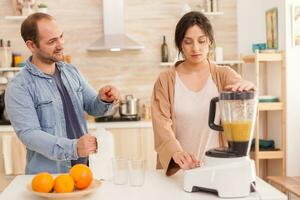 mulher mistura frutas dentro batido criador enquanto marido abre leite garrafa. saudável despreocupado e alegre estilo de vida, comendo dieta e preparando café da manhã dentro acolhedor ensolarado manhã foto