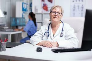 confiante com experiência Senior médico dentro branco casaco com estetoscópio que segurando vídeo conferência e consultando parceiros ou pacientes, olhando para dentro Câmera. médico aconselhamento paciente usando vídeo ligar foto