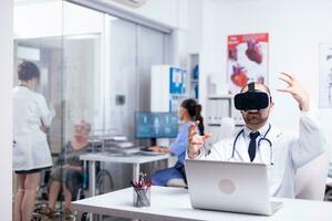 retrato do profissional caucasiano masculino médico dentro vr óculos sentado dentro hospital gabinete e fazer gestos usando virtual realidade inovações enquanto fêmea enfermeira trabalhando dentro fundo foto