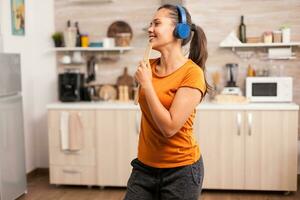 jovem mulher ouvindo música em fones de ouvido cantando em de madeira colher dentro cozinha. enérgico, positivo, feliz, engraçado e fofa dona de casa dançando sozinho dentro a casa. entretenimento e Leise sozinho às casa foto