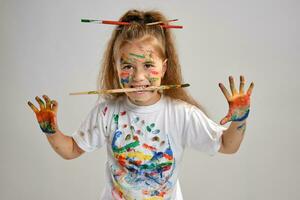 pequeno menina dentro branco camiseta, com escovas dentro dela cabelo é posando em pé isolado em branco, gesticulando com pintado mãos e face. fechar-se. foto