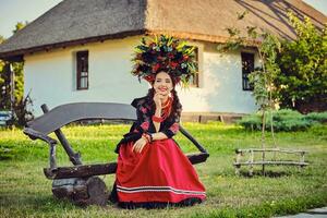 morena menina dentro uma branco ucraniano autêntico nacional traje e uma guirlanda do flores é posando sentado em uma de madeira Banco contra uma branco cabana. foto