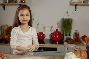 lindo pequeno menina vestido dentro uma branco blusa é fazer uma massa para cozimento uma pão às uma cozinha. foto