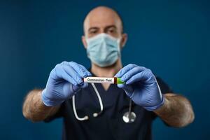 médico dentro mascarar, azul luvas, médico terno. segurando vermelho laboratório teste tubo com verde boné dentro mãos. azul fundo. coronavírus, COVID-19. fechar acima foto