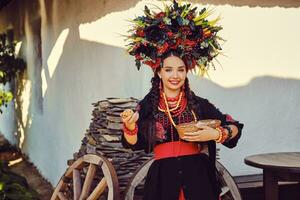 morena menina dentro Preto e vermelho bordado ucraniano autêntico nacional traje e uma guirlanda do flores é posando em pé contra uma branco cabana. foto