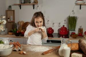 lindo pequeno menina vestido dentro uma branco blusa é fazer uma massa para cozimento uma pão às uma cozinha. foto