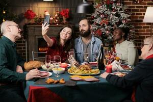 feliz pessoas leva fotos juntos às lar, desfrutando jantar festa com óculos do álcool cercado de natal decorações. diverso amigos e família levando As fotos durante Natal celebração.