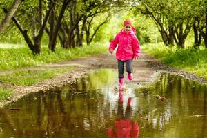 fofa pequeno menina vestindo chuva chuteiras pulando para dentro uma poça foto