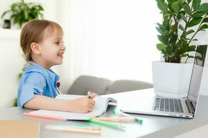 fofa pequeno menina é sentado às mesa com dela computador portátil e estudando conectados foto