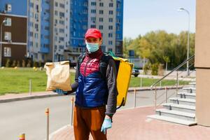 Comida Entrega homem dentro uma protetora mascarar e luvas com uma termo mochila perto uma casa durante a quarentena período foto