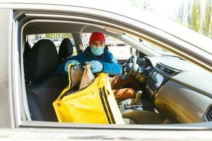 Comida Entrega homem dentro uma protetora mascarar e luvas com uma termo mochila perto uma carro durante a quarentena período foto