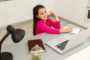 controlo remoto aulas. a criança sorrisos alegremente e pega conhecimento remotamente. pequeno menina estude conectados Aprendendo a partir de casa com computador portátil. conectados escola. foto