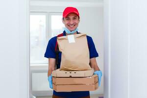 Entrega homem empregado dentro vermelho boné camiseta uniforme mascarar luvas dar Comida ordem pizza caixas isolado em amarelo fundo estúdio. serviço quarentena pandemia coronavírus vírus gripe 2019-ncov conceito foto