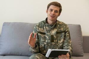 sorridente milenar homem dentro camuflar uniforme segurando moderno digital tábua, conversando com família, posando em branco estúdio fundo, cópia de espaço. moderno tecnologias e militares pessoal conceito. foto