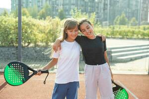 crianças e Esportes conceito. retrato do sorridente meninas posando ao ar livre em padel quadra com raquetes e tênis bolas foto