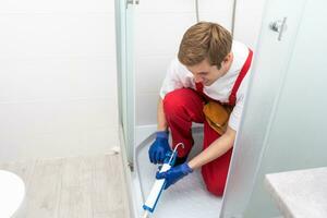 jovem homem dentro uniforme reparos a chuveiro porta dentro a banheiro. uma masculino reparador reparos a chuveiro cabine. foto