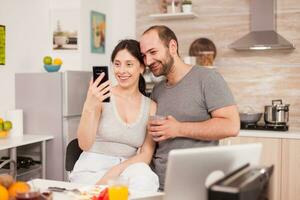 feliz jovem casal sorridente para selfie durante café da manhã. alegre casado marido e esposa fazer engraçado rostos enquanto levando uma foto durante café da manhã dentro cozinha.