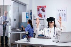 caucasiano fêmea médico dentro branco vestido e vr fone de ouvido sentado às escrivaninha dentro gabinete batendo e rolagem com mãos dentro ar. virtual realidade conceito. cuidados de saúde futurista. médico trabalhando dentro futuro. foto