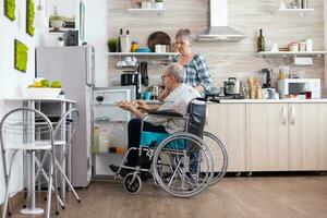 Desativado Senior masculino dentro cadeira de rodas levando ovos cartão a partir de geladeira ajudando dele esposa dentro cozinha. Senior mulher preparando café da manhã para deficiente marido, vivo com homem com caminhando deficiências foto