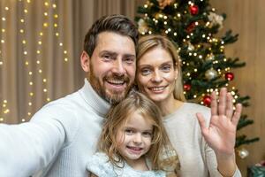 Natal família falando em telefone vídeo chamar, marido esposa e filha olhando às Smartphone Câmera e falando em telefone cumprimento amigos para Natal e Novo ano às casa perto Natal árvore. foto